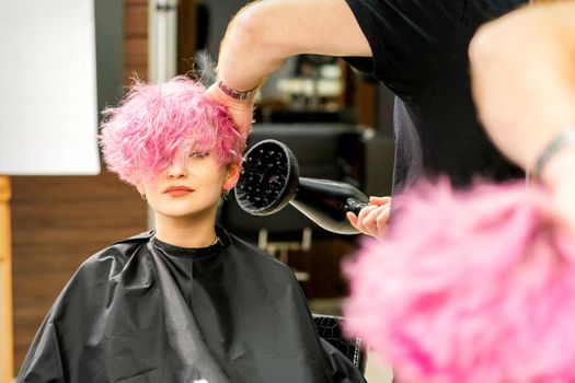 A male hairdresser professional drying stylish pink hair of the female client with a blow dryer in a beauty salon