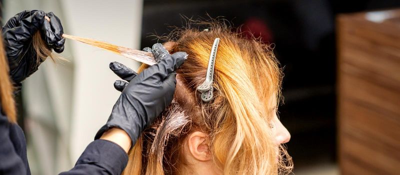 Closeup back view of hairdresser's hands in gloves applying dye to a strand of hair of redhead young woman in a hair salon