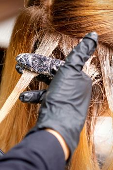 Closeup back view of hairdresser's hands in gloves applying dye to a strand of hair of redhead young woman in a hair salon
