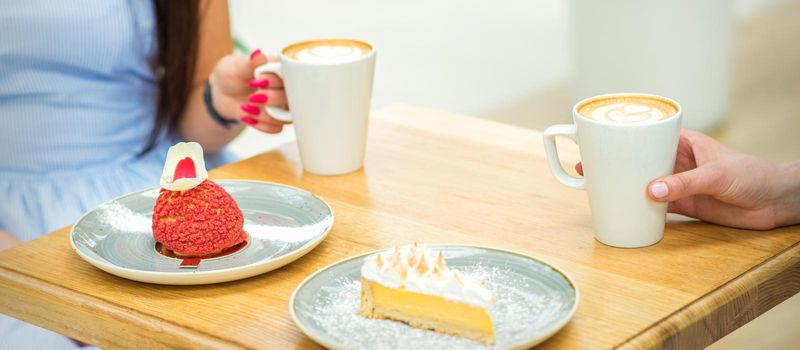 Two young women with cups of coffee and pieces of cake sitting at the table in a cafe outdoors