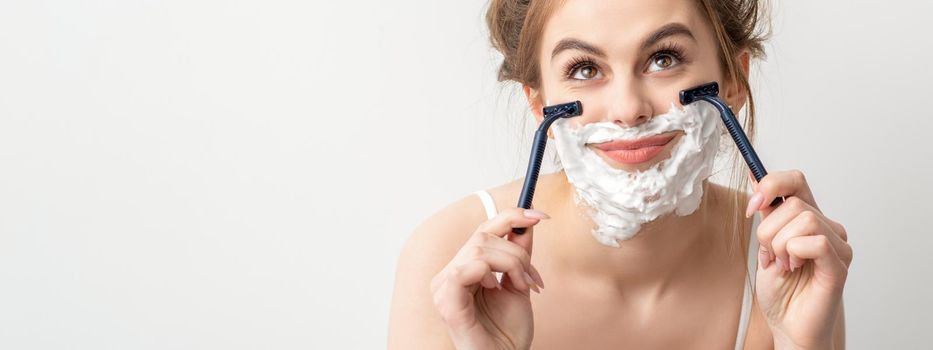 Beautiful young caucasian smiling woman shaving her face with razor looking up on white background