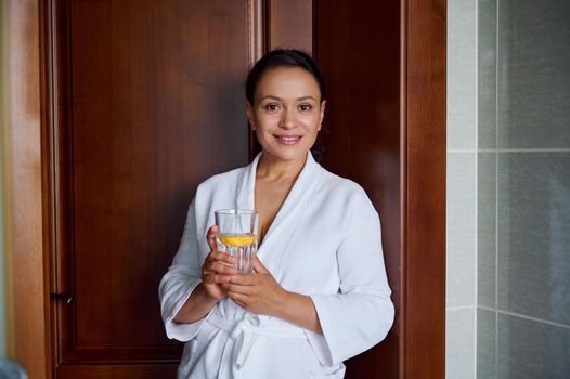Close-up portrait. Beautiful attractive Hispanic woman with glowing skin, in bathrobe, smiles looking at camera, holds glass with lemon water for good start to the day. Diet, vitality, healthy habits