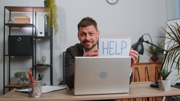Hopeless sad businessman in formal wear suit working on laptop computer, showing inscription note text Help. Overworked tired freelance business man indoors at office. Poverty, bankrupt, deadline