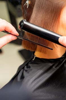 Hair stylist's hands straightening short hair of young brunette woman with flat iron and comb in a beauty salon