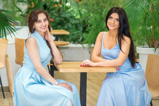Two beautiful smiling caucasian young women in long blue dresses sitting at the table and looking at the camera in a cafe outdoors