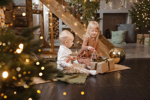 Little brother and sister play on Christmas eve in a beautiful house decorated for the New Year holidays. Children are playing with a Christmas gift. Scandinavian-style interior with live fir trees and a wooden staircase.