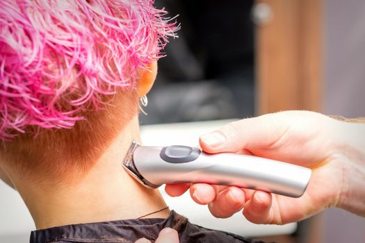 Back view of hairdresser's hand shaving nape and neck with electric trimmer of young caucasian woman with short pink hair in beauty salon