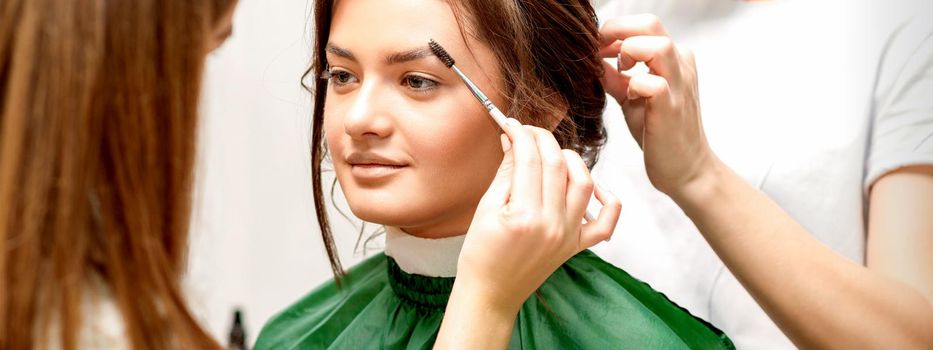 Makeup artist combing eyebrows and hairstylist preparing hairstyle for beautiful young caucasian bride in a beauty salon. Tandem of two beauty masters