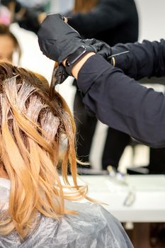 Back view of a female hairdresser in gloves coloring hair of redhead young woman with light or white dye in a beauty salon