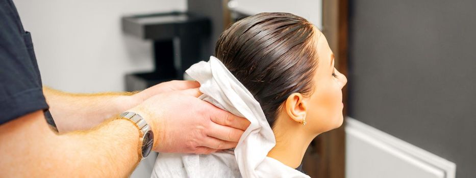 A male hairdresser's hands wipe the hair of a female client with a white towel after shampooing in a beauty salon