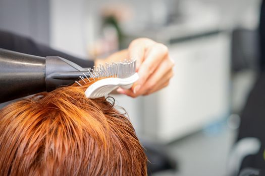 Closeup of master's hand with blow-drying and hairbrush blowing female red hair in a salon