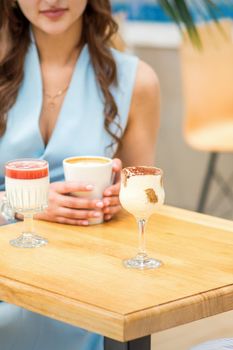 Hands of a young woman holding a cup of coffee with different cocktails on the table at the cafe