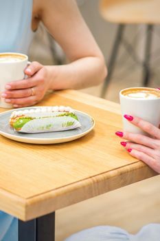 Two young women with cups of coffee and pieces of cake sitting at the table in a cafe outdoors