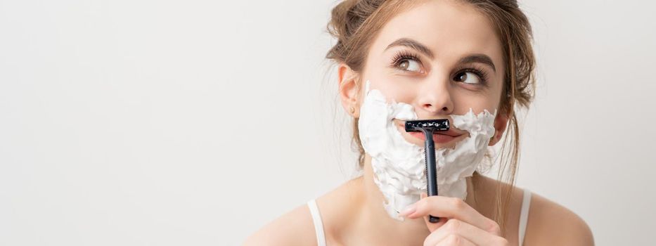 Beautiful young caucasian smiling woman shaving her face with razor looking up on white background