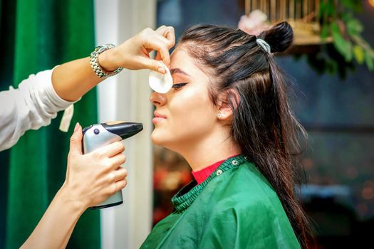 Side view of a make-up artist using aerograph making an airbrush makeup foundation on a female face in a beauty salon