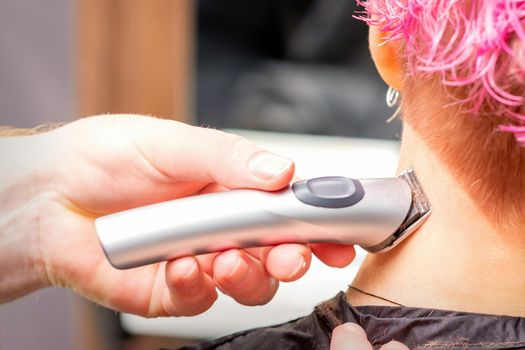 Back view of hairdresser's hand shaving nape and neck with electric trimmer of young caucasian woman with short pink hair in beauty salon