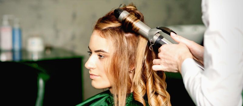 Hairdresser using curling tongs curls long brown hair on the young caucasian girl in a beauty salon
