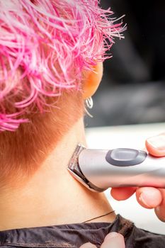 Back view of hairdresser's hand shaving nape and neck with electric trimmer of young caucasian woman with short pink hair in beauty salon