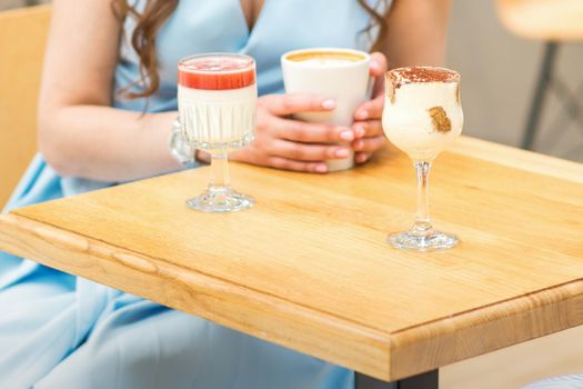 Hands of a young woman holding a cup of coffee with different cocktails on the table at the cafe