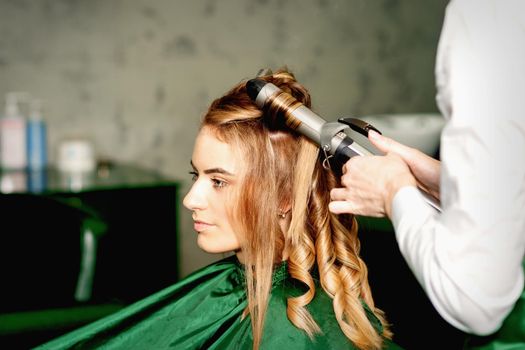 Hairdresser using curling tongs curls long brown hair on the young caucasian girl in a beauty salon