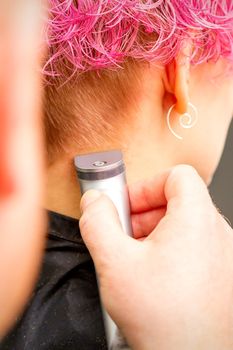 Back view of hairdresser's hand shaving nape and neck with electric trimmer of young caucasian woman with short pink hair in beauty salon