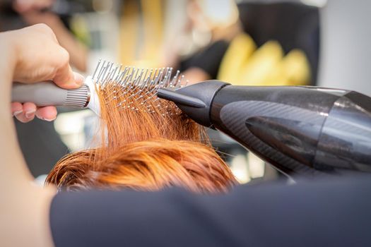 Closeup of master's hand with blow-drying and hairbrush blowing female red hair in a salon
