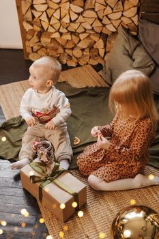 Little brother and sister play on Christmas eve in a beautiful house decorated for the New Year holidays. Children are playing with a Christmas gift. Scandinavian-style interior with live fir trees and a wooden staircase.