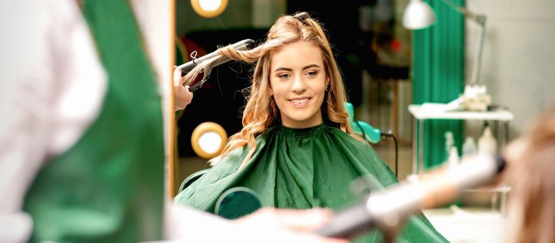 Hairdresser using curling tongs curls long brown hair on the young caucasian girl in a beauty salon