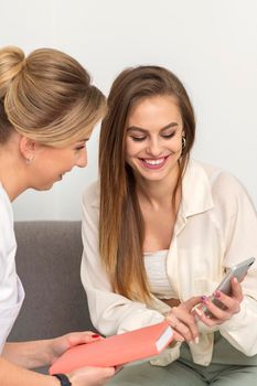 Two young women doctor with pacient smiling and looking at smartphone sitting in hospital office, consultation with a doctor