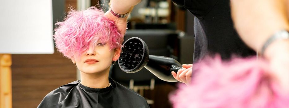 A male hairdresser professional drying stylish pink hair of the female client with a blow dryer in a beauty salon