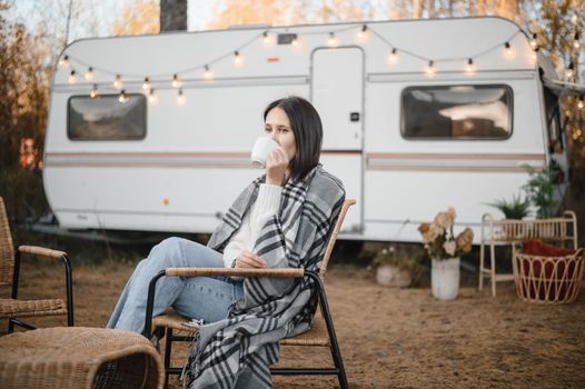 Caucasian woman in a knitted hat wrapped in a plaid and drinks a warming drink outdoors. Travel in a motor home in the fall