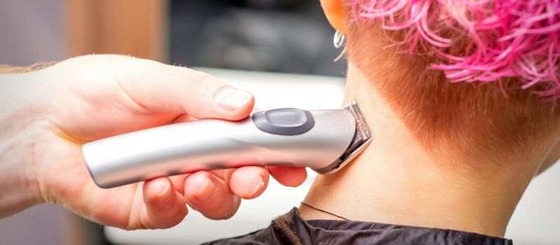 Back view of hairdresser's hand shaving nape and neck with electric trimmer of young caucasian woman with short pink hair in beauty salon