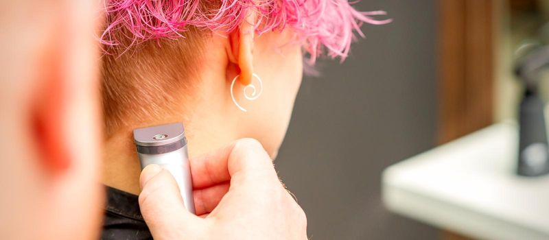 Back view of hairdresser's hand shaving nape and neck with electric trimmer of young caucasian woman with short pink hair in beauty salon