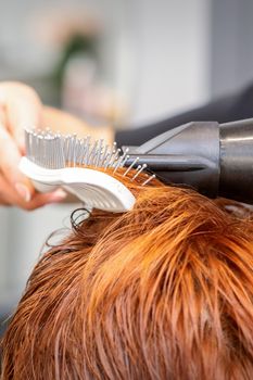 Closeup of master's hand with blow-drying and hairbrush blowing female red hair in a salon