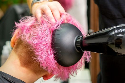 A male hairdresser professional drying stylish pink hair of the female client with a blow dryer in a beauty salon