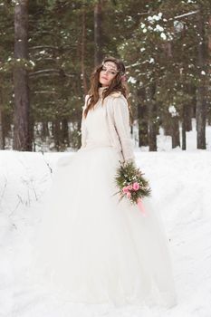 Beautiful bride in a white dress with a bouquet in a snow-covered winter forest. Portrait of the bride in nature.