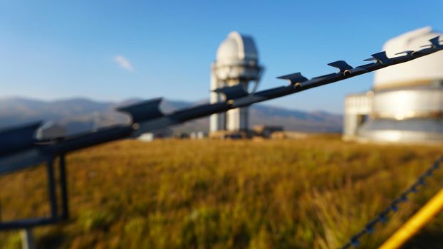 A large observatory is protected by a barbed fence. There is a long sharp wire around perimeter. Buildings with domes and a telescope are visible in distance. Yellow-green grass. Mountainous terrain