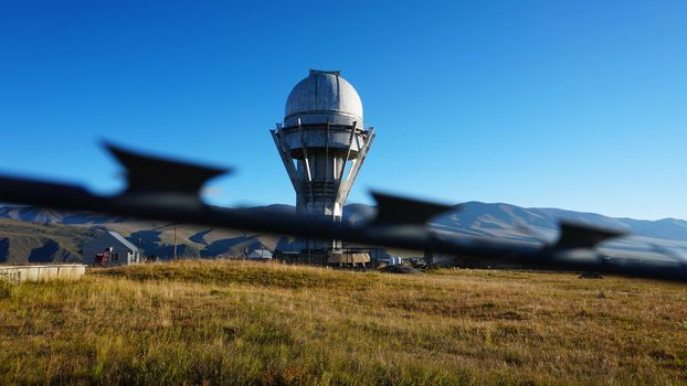 A large observatory is protected by a barbed fence. There is a long sharp wire around perimeter. Buildings with domes and a telescope are visible in distance. Yellow-green grass. Mountainous terrain