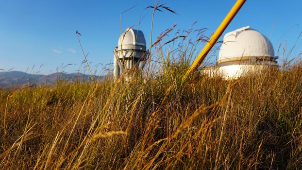 Large buildings of the observatory in the form of a dome. The telescopes are closed. Beautiful tall yellow-green grass develops in the wind. Blue sky and green hills in the distance. The Assy Plateau