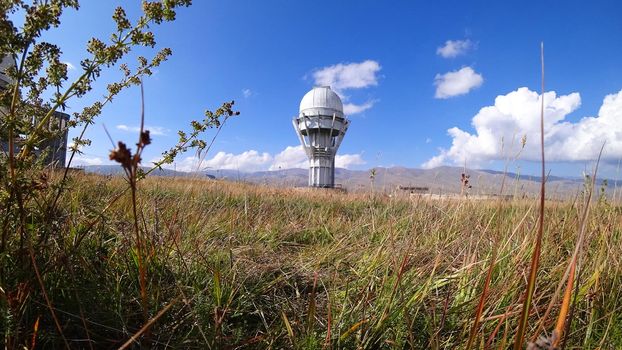 Large buildings of the observatory in the form of a dome. The telescopes are closed. Beautiful tall yellow-green grass develops in the wind. Blue sky and green hills in the distance. The Assy Plateau