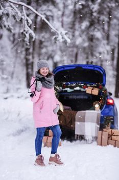A woman in a winter snow-covered forest in the trunk of a car decorated with Christmas decor. A female photographer holds a camera in her hands.