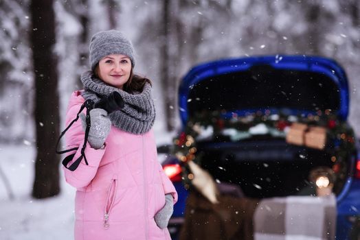 A woman in a winter snow-covered forest in the trunk of a car decorated with Christmas decor. A female photographer holds a camera in her hands.