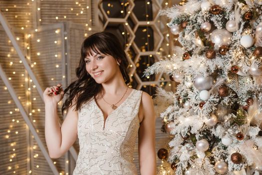 A brunette woman in a beautiful light tight dress next to a Christmas tree. The magical atmosphere of Christmas Eve on December 25. Decoration for the holiday in a light style with lots of lights and light bulbs.