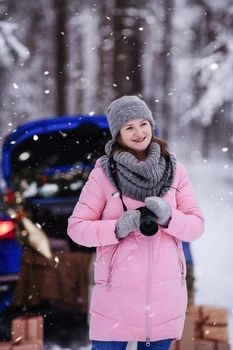A woman in a winter snow-covered forest in the trunk of a car decorated with Christmas decor. A female photographer holds a camera in her hands.