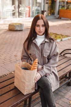 A stylish brunette woman walks around the autumn city. The brunette is sitting on a bench outside with a glass of coffee and a phone, holding a craft bag with a baguette.