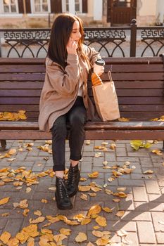 A stylish brunette woman walks around the autumn city. The brunette is sitting on a bench outside with a glass of coffee and a phone, holding a craft bag with a baguette.