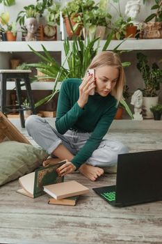 A blonde woman in a room with a lot of green indoor plants is working on a laptop. The concept of biophysical design in the interior. Work from home, work as a freelancer.