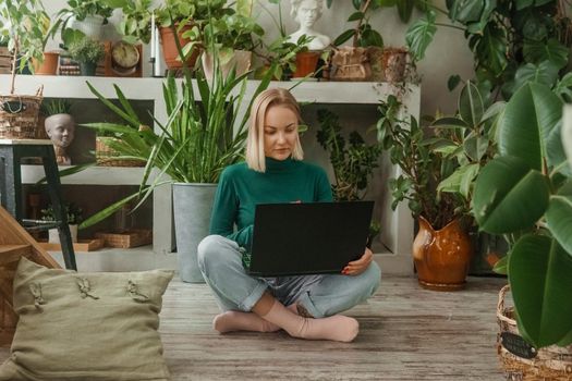 A blonde woman in a room with a lot of green indoor plants is working on a laptop. The concept of biophysical design in the interior. Work from home, work as a freelancer.