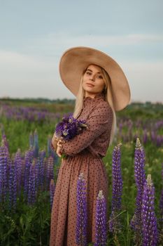 A beautiful woman in a straw hat walks in a field with purple flowers. A walk in nature in the lupin field.