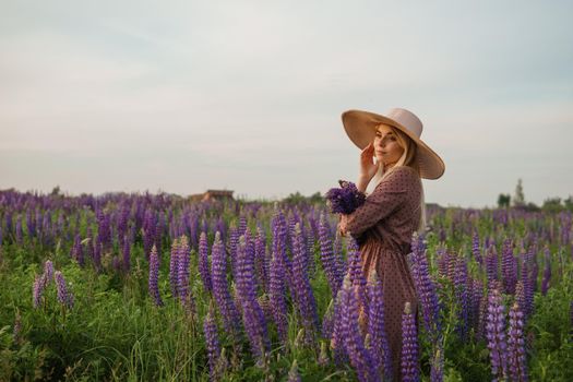 A beautiful woman in a straw hat walks in a field with purple flowers. A walk in nature in the lupin field.